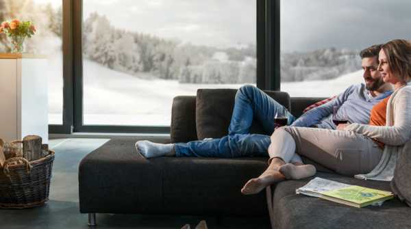 Personen sitzen entspannt auf einem modernen, dunklen Sofa vor einem knisternden Kaminfeuer, mit Blick auf eine verschneite Landschaft durch grosse Panoramafenster. Ein Korb mit Holz steht neben dem Kamin, Zeitschriften und Schuhe liegen im Wohnbereich.