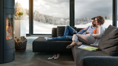 Personen sitzen entspannt auf einem modernen, dunklen Sofa vor einem knisternden Kaminfeuer, mit Blick auf eine verschneite Landschaft durch grosse Panoramafenster. Ein Korb mit Holz steht neben dem Kamin, Zeitschriften und Schuhe liegen im Wohnbereich.