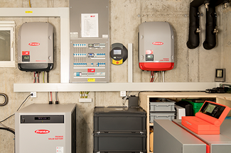 Technikraum mit mehreren an der Betonwand montierten Fronius‑Wechselrichtern, einem Elektro‑Verteilerkasten mit Sicherungen, einem Batteriespeicher sowie sauber verlegten Kabeln und Leitungen; im Vordergrund befinden sich zusätzliche Geräte und Aufbewahrungsboxen.